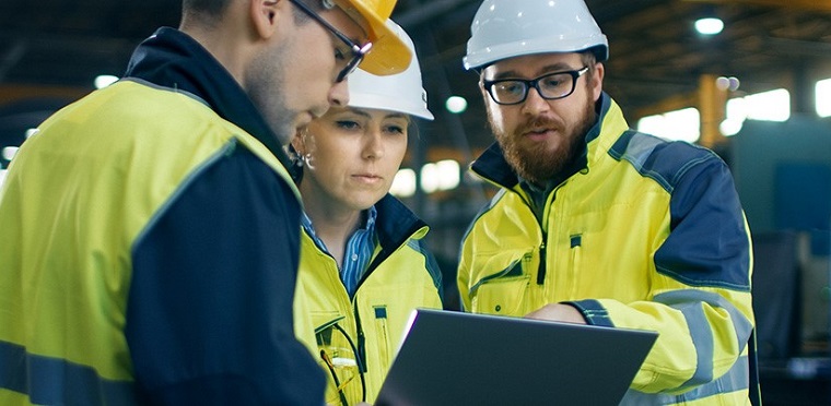Three workers in hard hats in a large building looking at a laptop